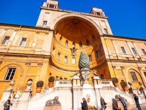 The image shows the Courtyard of the Pinecone at one of the top 10 museums, the Vatican Museums in Vatican City. A large bronze pinecone sculpture stands center stage before a semicircular niche, surrounded by ornate architecture with columns, statues, and a curved balustrade under a clear blue sky.